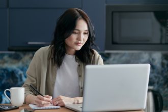 A woman wearing a beige button-up shirt looks at her laptop and takes notes at a counter.