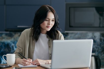 A woman wearing a beige button-up shirt looks at her laptop and takes notes at a counter.
