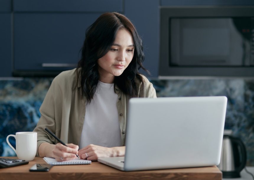 A woman wearing a beige button-up shirt looks at her laptop and takes notes at a counter.